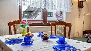 A dining table set with blue plates and cups, a vase with flowers, and a window with lace curtains. - Apartment 'Ferienwohnung Auf Der Insel Rügen' with Shared Terrace and Shared Garden (Bergen auf Rügen)