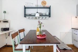 A dining area with a wooden table, chairs, a bowl of fruit, a vase with flowers, a radio, and a shelf with dishes and glasses.