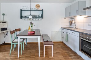 A modern kitchen with a wooden table, chairs, a bench, a hanging light, a clock, and a built-in oven.