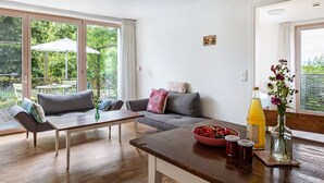 A modern living room with a sofa, coffee table, and dining table. There is a large window with a view of greenery outside. - Apartment 'Schambachhof' (Bötzingen am Kaiserstuhl)