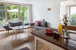 A modern living room with a sofa, coffee table, and dining table. There is a large window with a view of greenery outside.