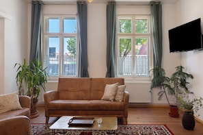 A living room with a brown sofa, a coffee table, a TV mounted on the wall, and large windows with curtains.