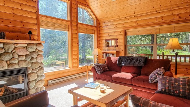 A log cabin interior with a stone fireplace, a red sofa, a wooden coffee table, and large windows offering a view of the outdoors. - Woodland Retreat: Cozy Cabin Near Trails & Mazama (Winthrop)