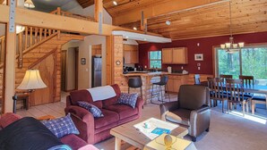 A spacious living area with a high ceiling, wooden beams, a kitchen with wooden cabinets, a dining area with a table and chairs, and a staircase leading to an upper level. - Woodland Retreat: Cozy Cabin Near Trails & Mazama (Winthrop)