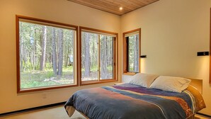 A bedroom with a large bed, three windows, and a view of a forest. - Petrichor - Mazama Modern Cabin (Mazama)