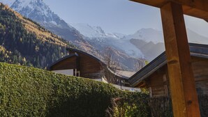 A wooden cabin with a curved roof, surrounded by a well-maintained hedge, set against a backdrop of snow-capped mountains and a clear sky.