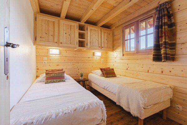 A wooden cabin room with two beds, a window with curtains, and built-in wooden cabinets.