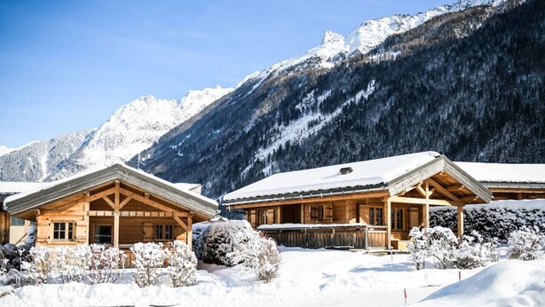 A wooden lodge with a covered porch and snow-covered surroundings, set against a backdrop of mountains.