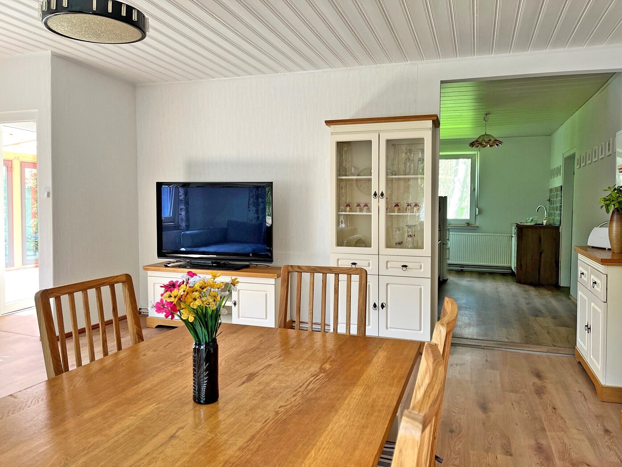 Een eetgedeelte met een houten tafel, stoelen, een bloevas, een televisie en een kast met glazen deuren.