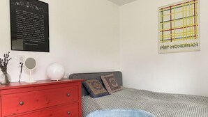 A bedroom with a red dresser, a round mirror, a white lamp, a patterned bedspread, and a framed artwork on the wall. - Süßes Gästehäuschen in 1A Lage (Bochum)