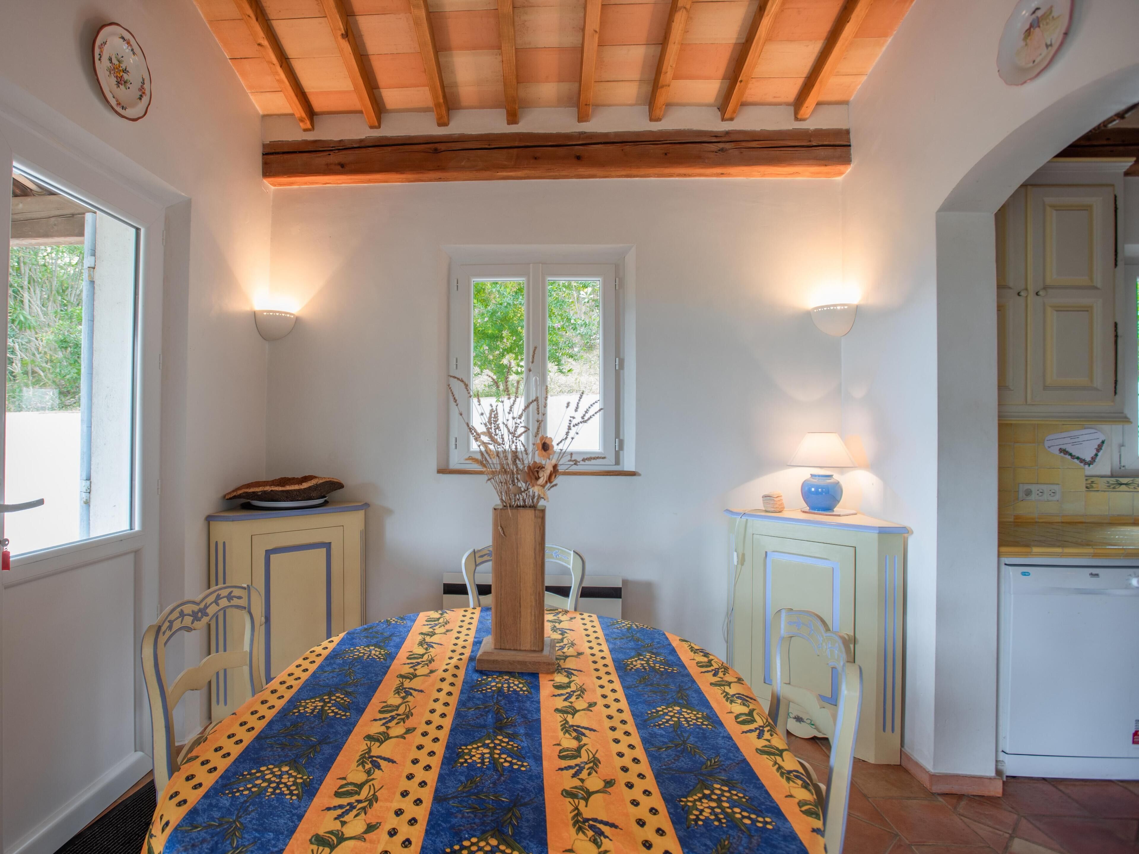 Un coin repas avec une table recouverte d’une nappe à motifs, un vase contenant des fleurs séchées et une fenêtre donnant sur un paysage verdoyant.