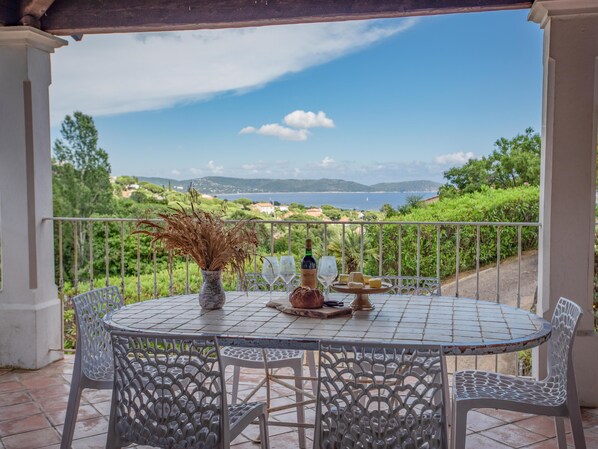 A round table with chairs on a balcony overlooking a scenic view of the sea and hills.
