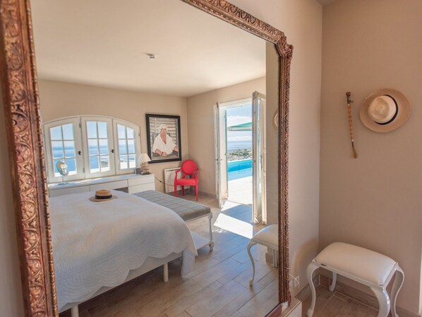 A bedroom with a bed, a red chair, a white bench, a large mirror, and a view of the ocean through an open door.