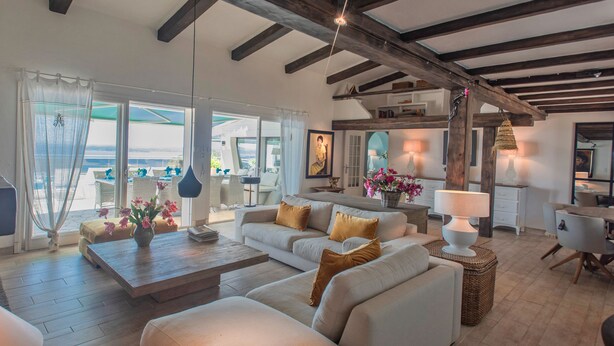 A spacious living area with a large sofa, a coffee table, and a dining table. There is a view of the outdoors through large windows.