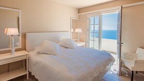 A bedroom with a large bed, two bedside tables with lamps, a chair, and a sliding glass door leading to a balcony with a view of the sea.