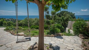 A stone patio with a tree in the center, surrounded by greenery and a view of the sea.