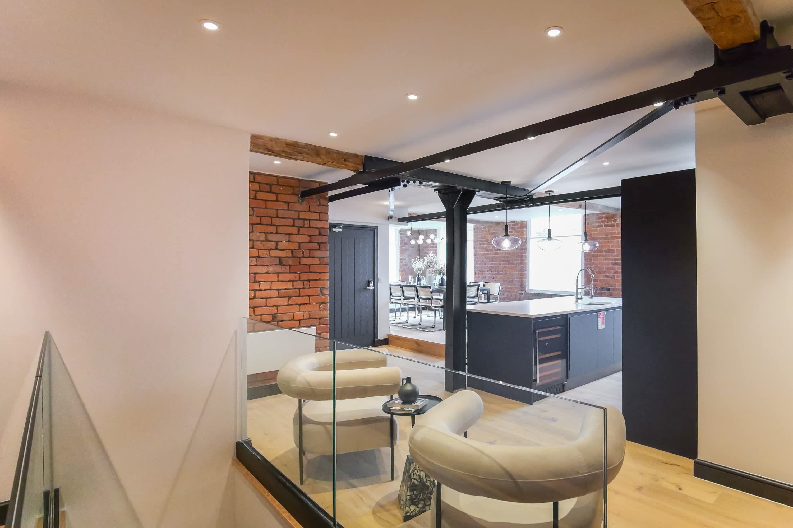 Un intérieur moderne avec une balustrade en verre, des fauteuils en cuir, une petite table et un coin cuisine visible à travers une porte ouverte.