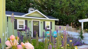 A quaint house with a green exterior and a dark brown door, surrounded by a variety of flowering plants and a white picket fence. - 🌹Cutest Garden Cottage🌹Lopez Village-Walk to Beach, Dining, Brewery, Farm Mkt
 (Lopez Island)