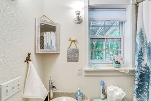 Ett badrum med en vit, strukturerad vägg, en upphängd handduk, en spegel med en dekorativ ram, ett fönster med utsikt över träd och en hylla med en vas med blommor.