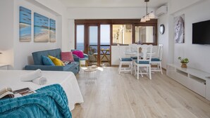 Una sala de estar moderna con un sofá, una mesa de comedor con sillas, un televisor montado en la pared y vistas al mar a través de amplias ventanas.