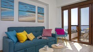 Un salón moderno con un sofá azul turquesa, cojines coloridos, una mesa de centro y un balcón con vistas al mar.