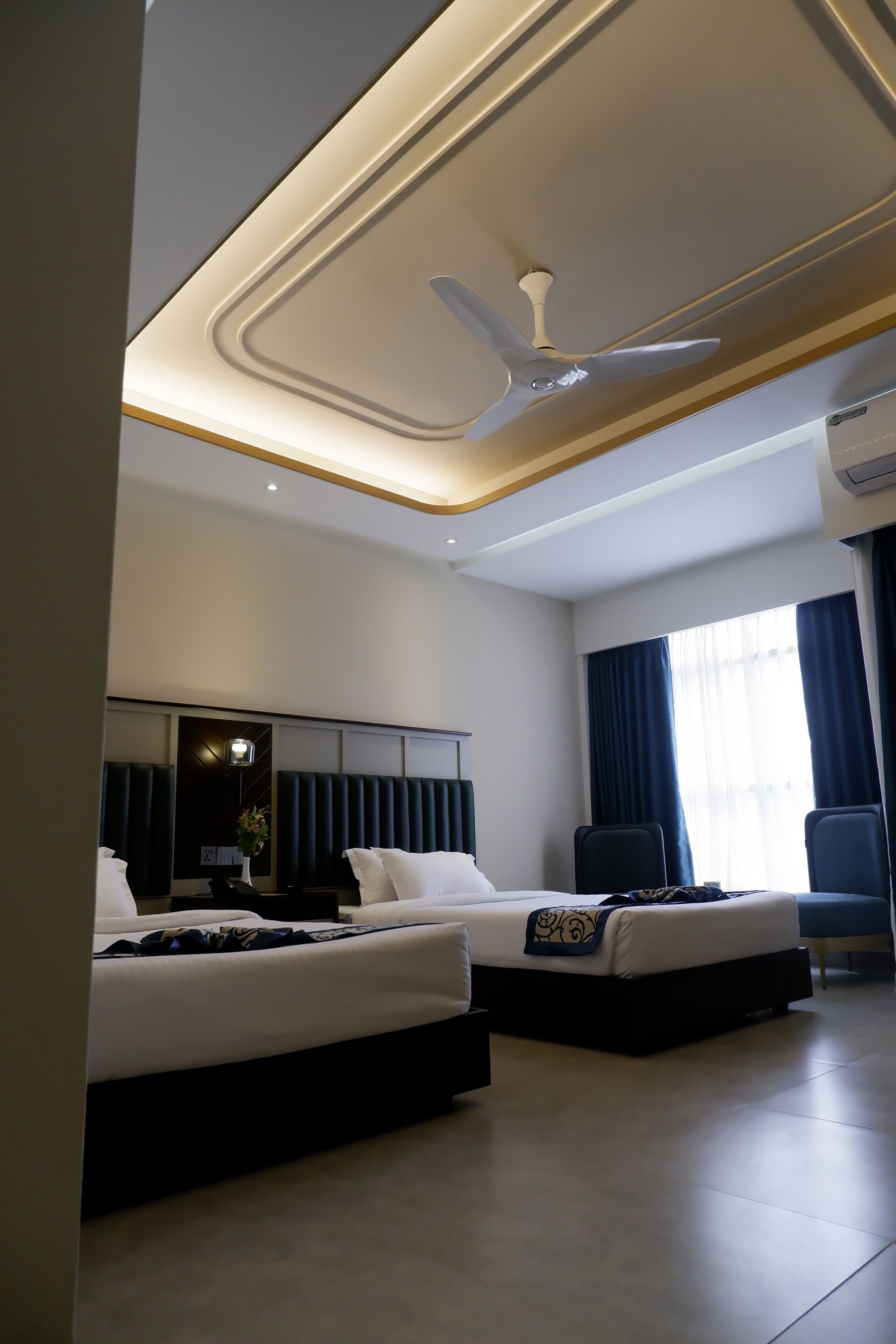 A modern hotel room with two beds, a ceiling fan, and a large window with curtains.