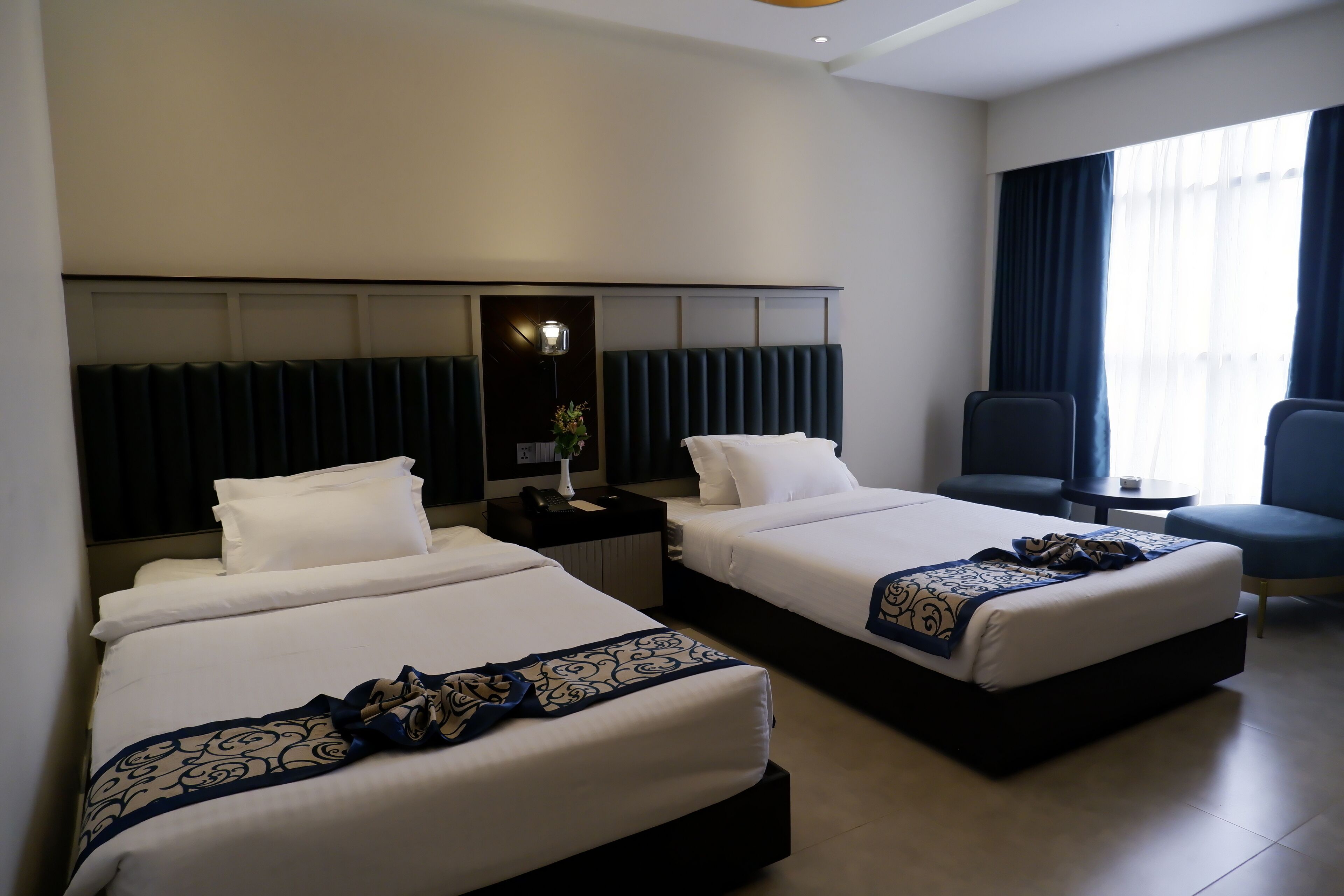 Habitación de hotel con dos camas, una mesita de noche con lámpara, una silla y vista al exterior por una ventana.