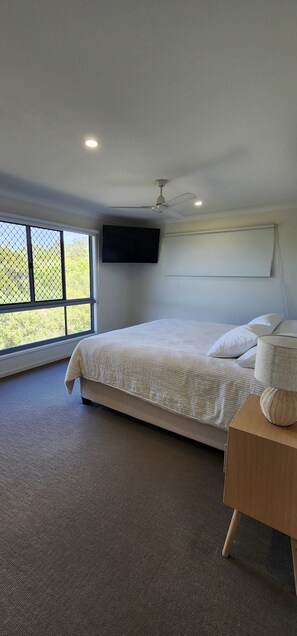 A bedroom with a bed, a ceiling fan, a TV, and a large window. - Starfish Beach House (Agnes Water)