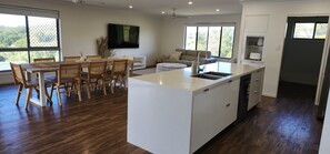 A modern kitchen with a central island, dining area with wooden chairs, and large windows. - Starfish Beach House (Agnes Water)