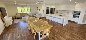 A modern kitchen with white cabinets, a central island with stools, and a dining area with a wooden table and chairs. - Starfish Beach House (Agnes Water)
