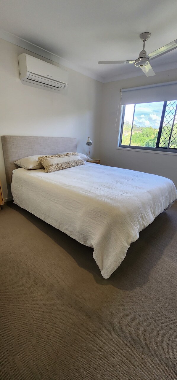A bedroom with a large bed, an air conditioning unit, a ceiling fan, and a window with a view of greenery. - Starfish Beach House (Agnes Water)