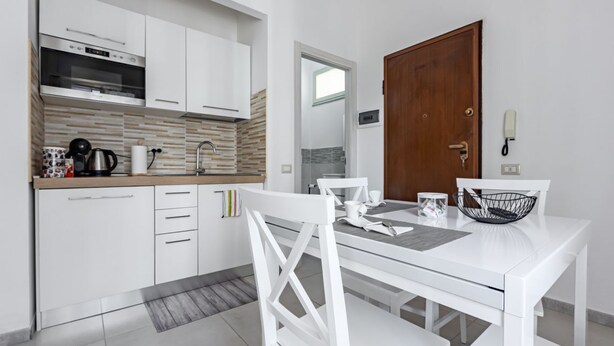 A modern kitchen with white cabinetry, a built-in oven, and a dining area with a white table and chairs. - GuestHost - Cozy apartment of approximately 28 sqm, ideal for 2 people, located on the ground floor of a building WITHOUT a lift (NOT suitable for disabled people).The apartment is located in a central neighborhood, served by all the main services: bars, (Cagliari)