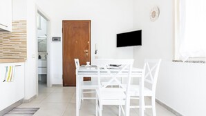 A modern kitchen with white cabinetry, a wooden door, a flat-screen TV mounted on the wall, and a dining area with white chairs and a table. - GuestHost - Cozy apartment of approximately 28 sqm, ideal for 2 people, located on the ground floor of a building WITHOUT a lift (NOT suitable for disabled people).The apartment is located in a central neighborhood, served by all the main services: bars, (Cagliari)