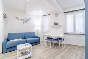 A modern living room with a blue sofa, a white coffee table, a dining table with chairs, a lamp, and a wall-mounted clock.