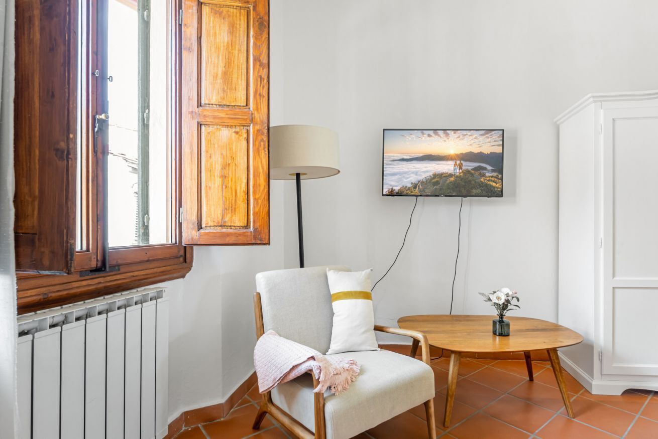A room with a wooden-framed window, a chair with a cushion, a table with a vase, and a wall-mounted TV displaying a sunset scene.