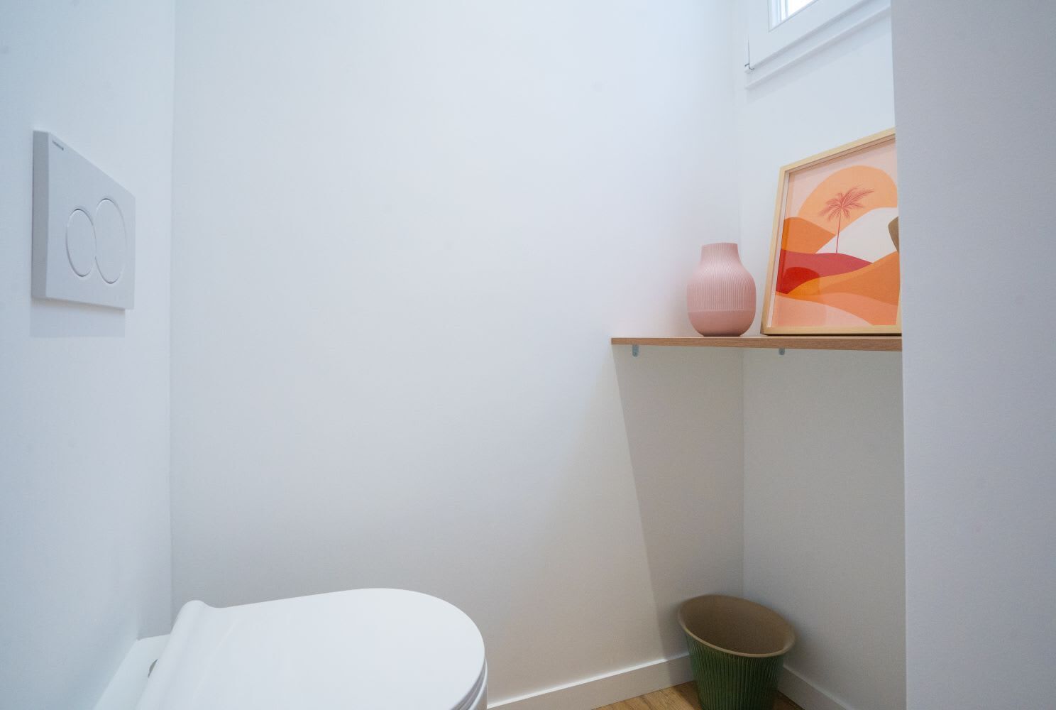 Un baño pequeño y limpio con un inodoro, un bote de basura, un estante con un florero y un cuadro enmarcado.