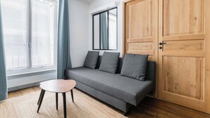 Une pièce avec un canapé gris, une petite table en bois, une grande porte en bois et une fenêtre avec des rideaux.