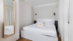 Un lit blanc avec une tête de lit en bois, un miroir et un mur décoré d’un motif.