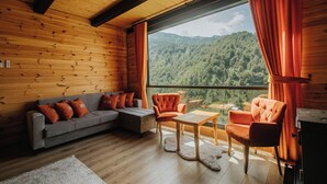 A cozy cabin interior with a wooden ceiling, a gray sofa with orange cushions, a wooden table, and a view of a forested mountain. - Ena Bungalov (Çamlihemsin)