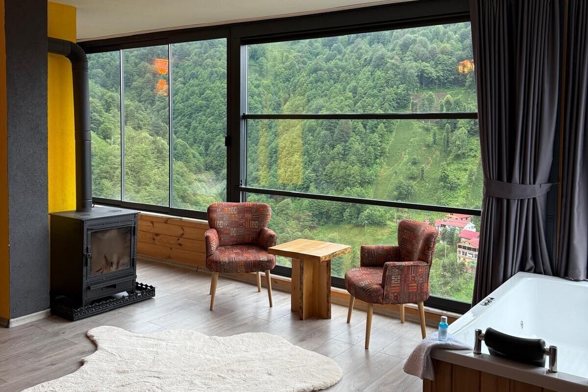 Una habitación moderna con un ventanal grande, un hogar a leña, dos sillones estampados, una pequeña mesa de madera y una bañera blanca.