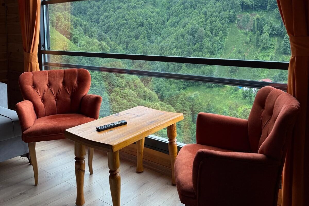 Una habitación con un gran ventanal que ofrece vistas a un valle boscoso. Hay dos sillones rojos y una mesa de madera con un control remoto.