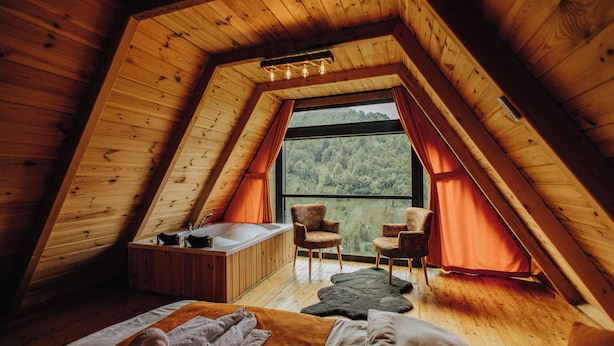 A cozy attic bedroom with a sloped ceiling, a large window with curtains, a bathtub, and two chairs. - Ena Bungalov (Çamlihemsin)