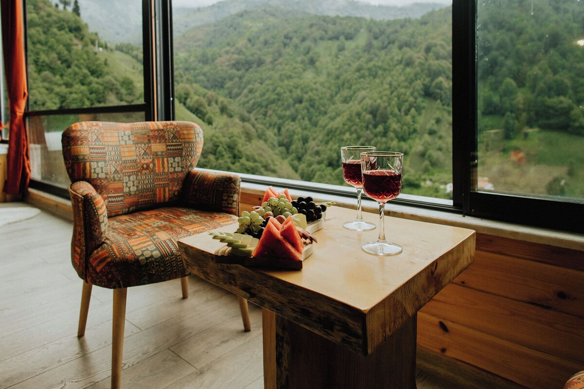 Una habitación con un ventanal grande, una mesa de madera, un sillón estampado y dos copas de vino.