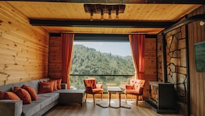 A cozy cabin interior with a wooden interior, a stone wall, a fireplace, and a view of a forested hillside. - Ena Bungalov (Çamlihemsin)