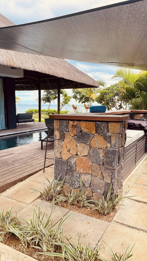 A stone pool with a thatched roof seating area, a stone wall with a bench, and a view of the sea.