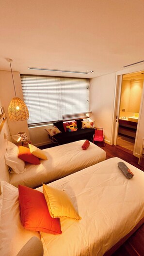 A hotel room with two beds, a sofa, a lamp, and a window with blinds.