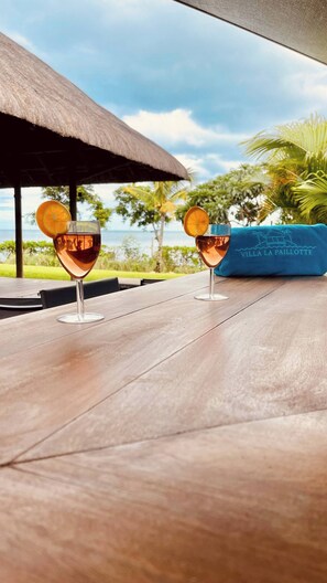 Two glasses of a pink beverage with orange slices on a wooden table, a thatched roof, and a view of the sea.