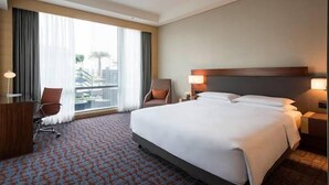 A hotel room with a large bed, a desk, a chair, and a view of the city through large windows. - Hotel Rajvanshi (Gurugram)