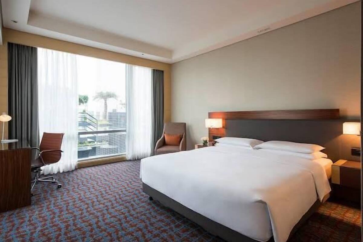 A hotel room with a large bed, a desk, a chair, and a view of the city through large windows.