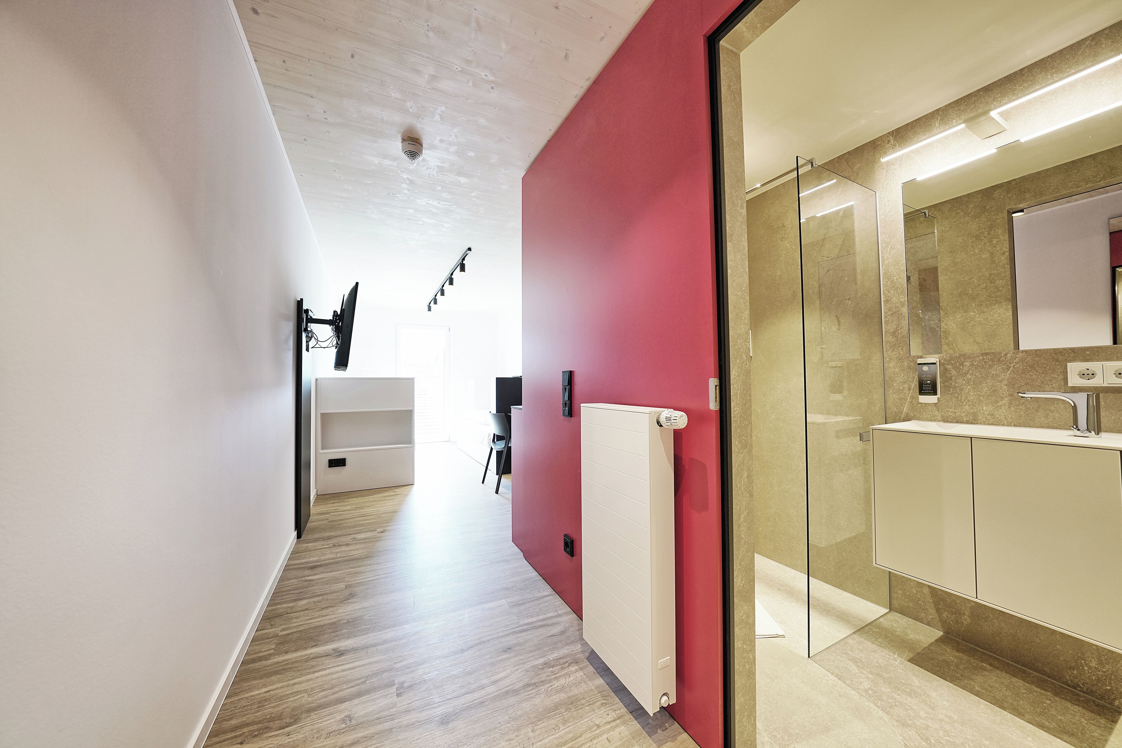 Een moderne hal met een rode accentmuur, een glazen douchecabine en een witte radiator.